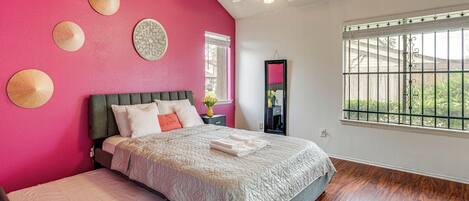 A bedroom with a large bed, a ceiling fan, a window with a view of greenery, and a pink wall.