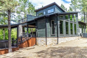 A two-story wooden cabin with a stone foundation, a wooden deck, and a staircase leading to a balcony. - ‘Sip on Inn’ Sips, Soaks & Fun Vibes in Hochatown! (Broken Bow)