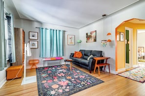 A living room with a black leather sofa, a wooden coffee table, and a colorful floral rug. There is a small orange stool and a white standing fan. - Families Welcome! Colorful Berkley Home w/ Yard (Berkley)