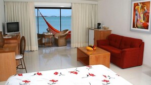 Camera d'albergo con un letto, un divano rosso, un tavolino in legno con della frutta, una TV, una scrivania con una sedia, un piccolo tavolo con una sedia e vista sul mare attraverso ampie finestre.