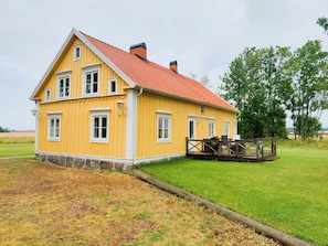 A yellow wooden house with a red roof, a wooden deck, and a grassy yard.