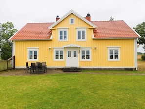 A yellow two-story house with a red-tiled roof, a white door, and a small porch with chairs.