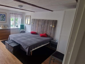 A bedroom with a wooden headboard, a bed with grey bedding, two red pillows, a bedside table, and a small stool.