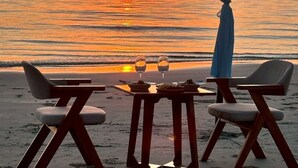 Un'area ristorazione sulla spiaggia, con tavolo e sedie sotto una tettoia, affacciata sull'oceano al tramonto.