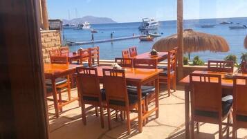 An outdoor seating area with wooden tables and chairs, a thatched umbrella, and a view of boats on the water.
