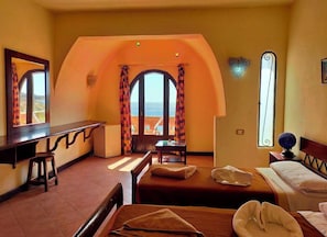 Een hotelkamer met een bed, een bureau, een stoel en uitzicht op de zee door een rondbogig raam.