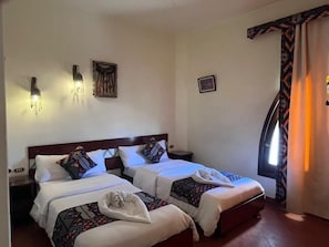 Habitación de hotel con dos camas, luces fijadas a la pared, un cuadro enmarcado y una ventana con cortinas.