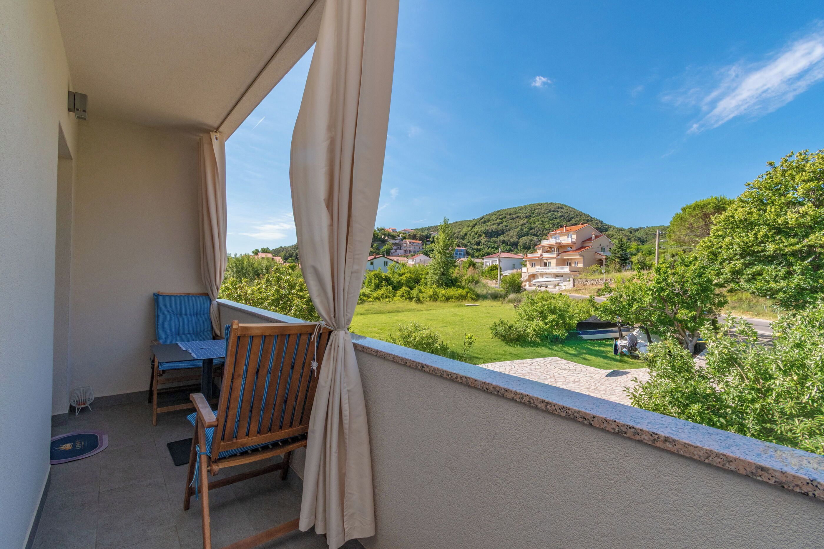 Un balcon avec une table et une chaise, une vue sur un paysage verdoyant et des maisons, et un ciel dégagé.