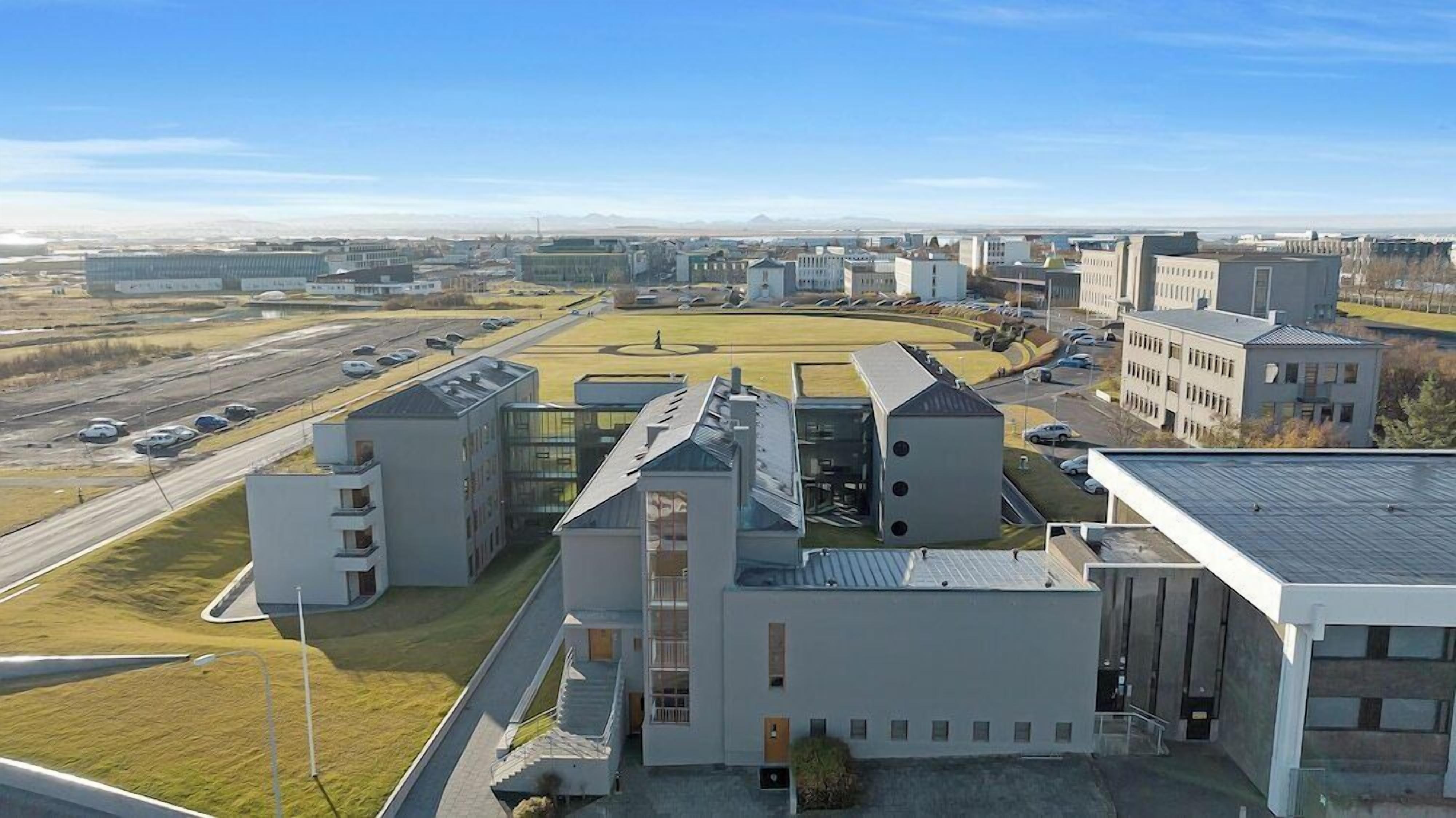 Un complejo moderno de edificios, con varias estructuras, incluyendo un edificio central de techo inclinado y un edificio adyacente más pequeño. Hay un estacionamiento con varios coches y un amplio espacio abierto al fondo.