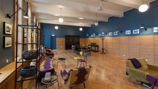 A spacious lobby with a wooden floor, a seating area with colorful chairs, a bookshelf, and framed pictures on the walls. - Hotel Gardur (Reykjavik)