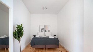 Un dormitorio con piso de madera, una cama con manta gris, dos almohadas, una mesita de noche con una lámpara y una planta en maceta.