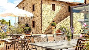 Un bâtiment en pierre avec un escalier, un espace extérieur aménagé avec des chaises et des tables en métal, et un grand parasol.