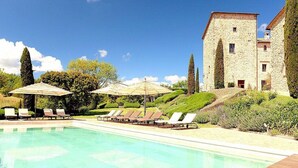 Une piscine entourée de verdure et d’un bâtiment en pierre, avec des chaises longues et des parasols à proximité.