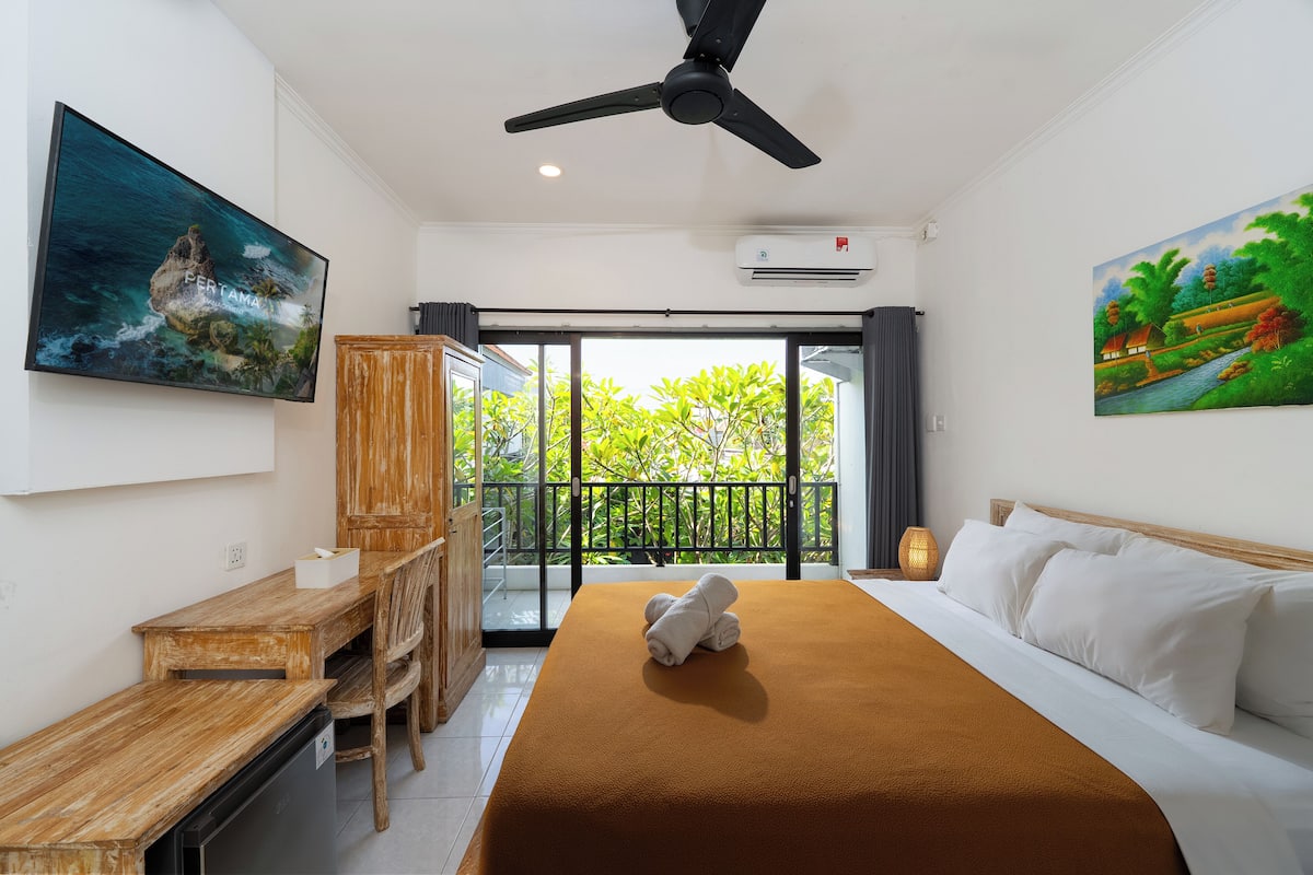 Une chambre d’hôtel équipée d’un lit, d’un bureau, d’une chaise, d’un ventilateur de plafond et d’un balcon offrant une vue sur la verdure.