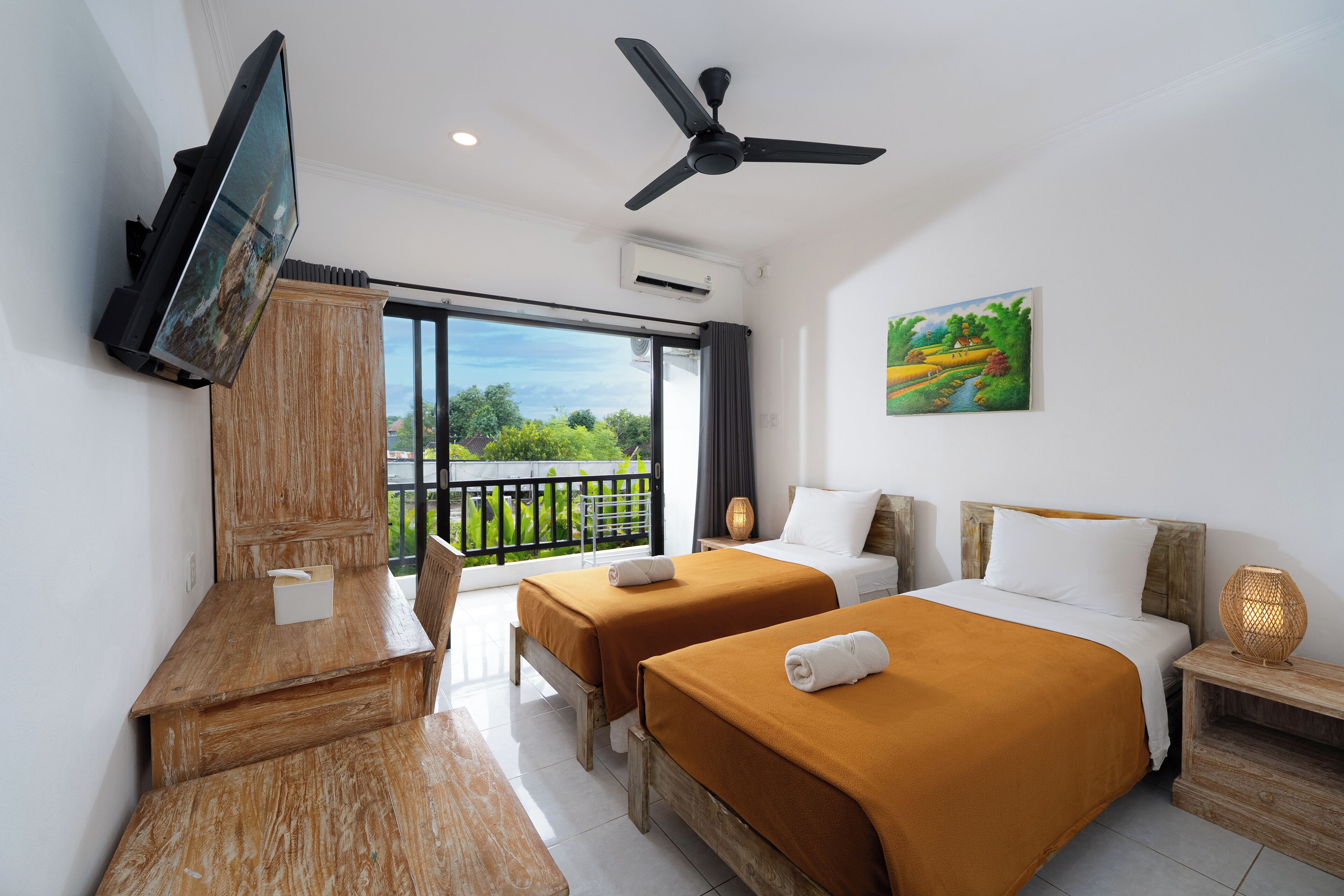 Habitación de hotel con dos camas, televisor, ventilador de techo y balcón con vistas a zonas verdes.