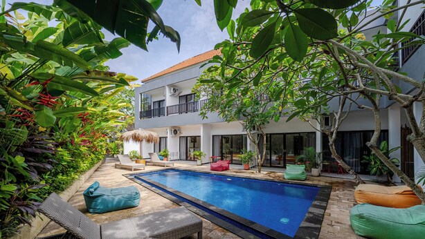 A swimming pool with lounge chairs and bean bags, surrounded by lush greenery and a modern building with balconies. - Uluwatu Surf Paradise (Ungasan)