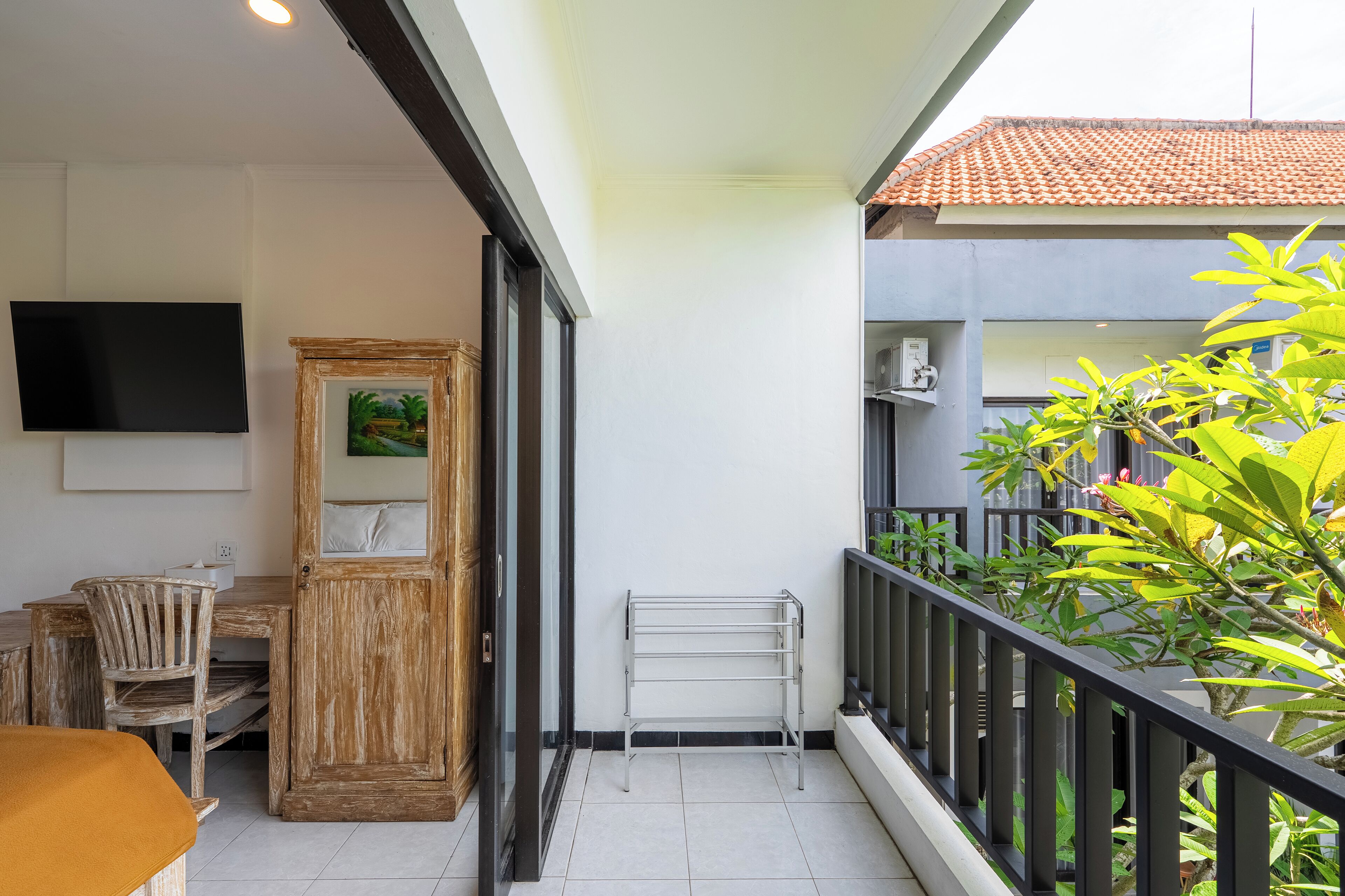 A balcony with a metal drying rack, a wooden cabinet with a painting, a TV mounted on the wall, and a chair.
