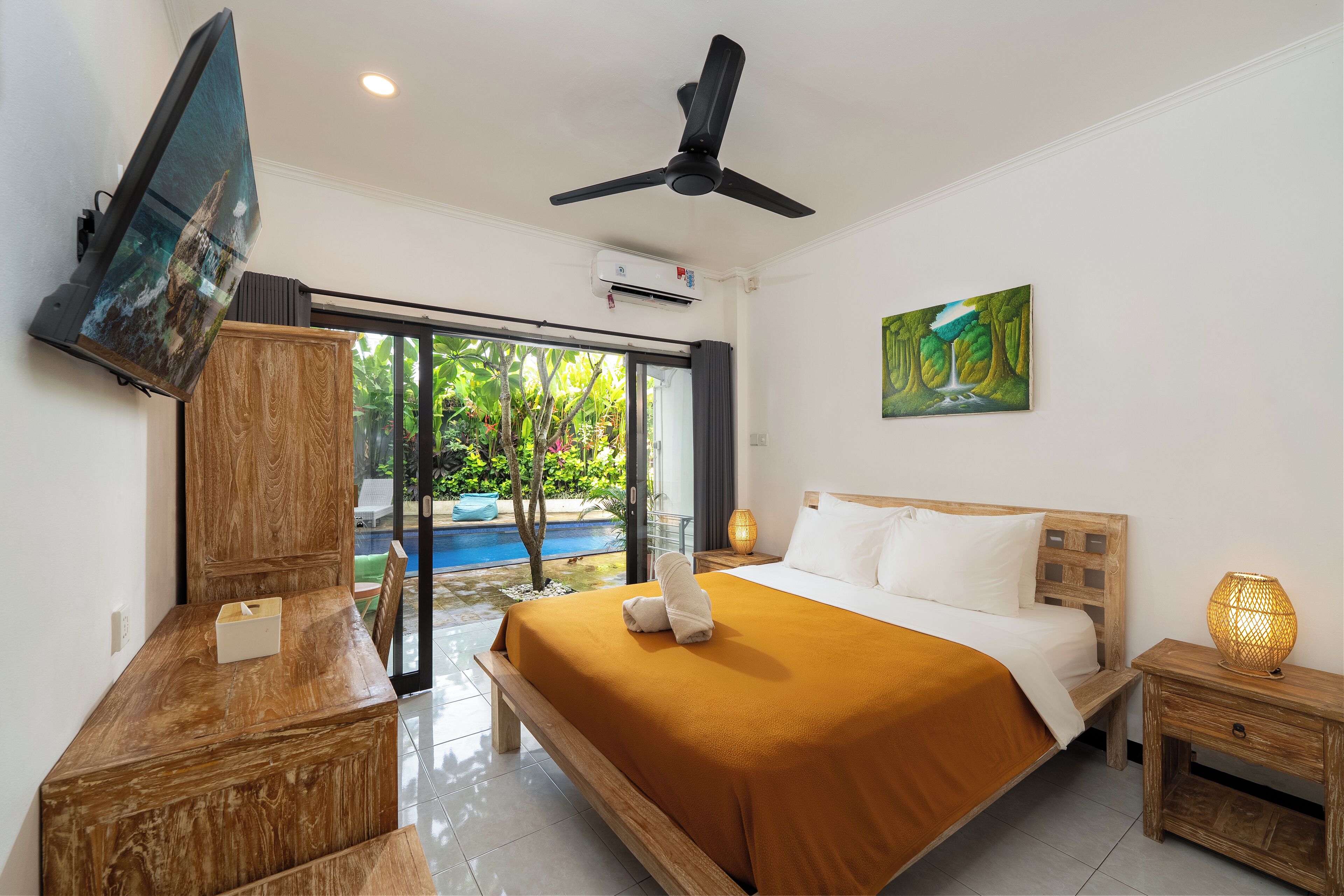 Habitación de hotel con cama, televisión, ventilador de techo, un cuadro y vista a una piscina.