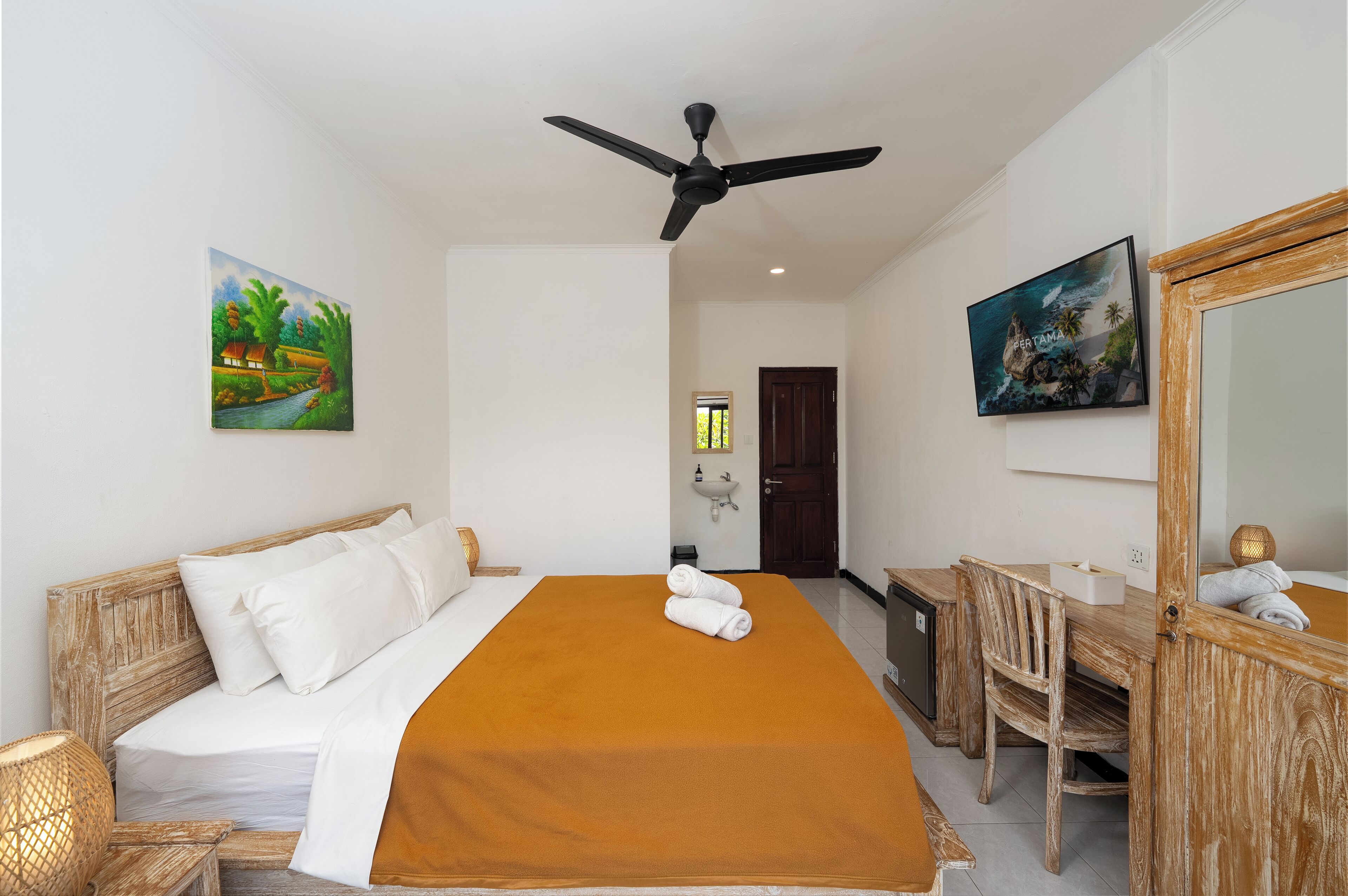 Quarto de hotel com cama, escrivaninha, cadeira, ventilador de teto e duas pinturas nas paredes.