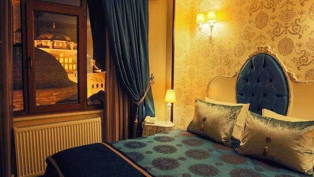 Une chambre d’hôtel avec un lit, une table de chevet, une lampe, une fenêtre donnant sur un immeuble et un papier peint à motifs.