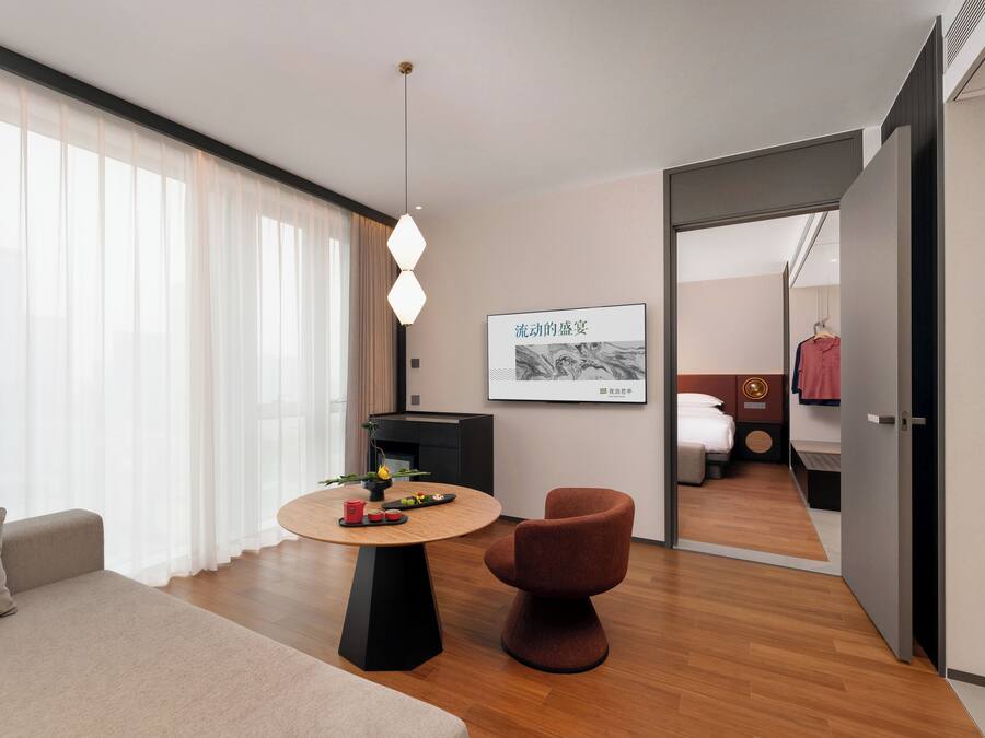 Una habitación de hotel moderna con una mesa redonda de madera, un sofá cómodo, una silla roja, una cama y un armario.