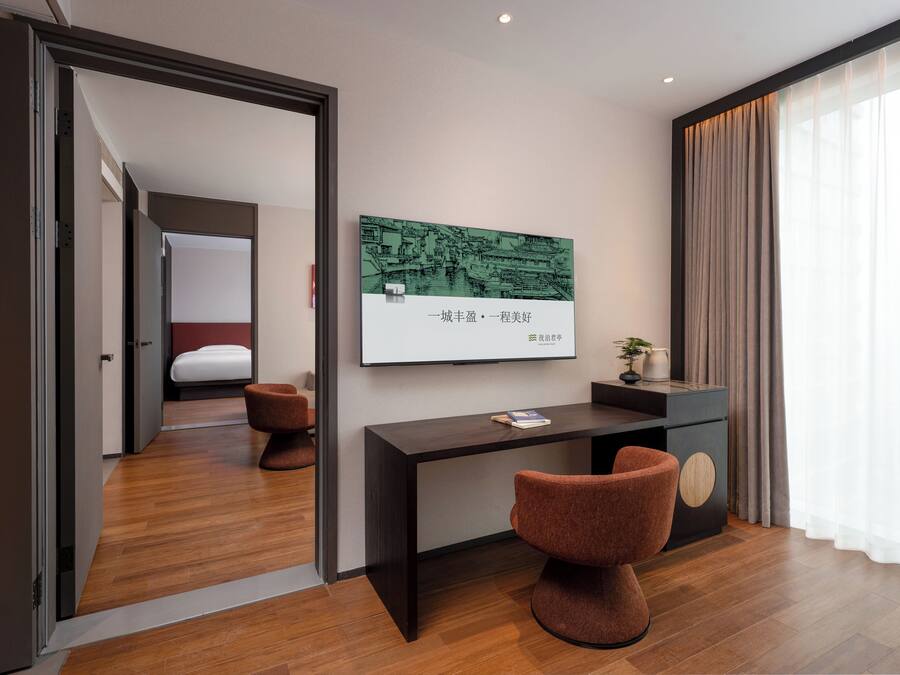 A modern hotel room with a wooden floor, a desk with a chair, a bed, and a large window with curtains.