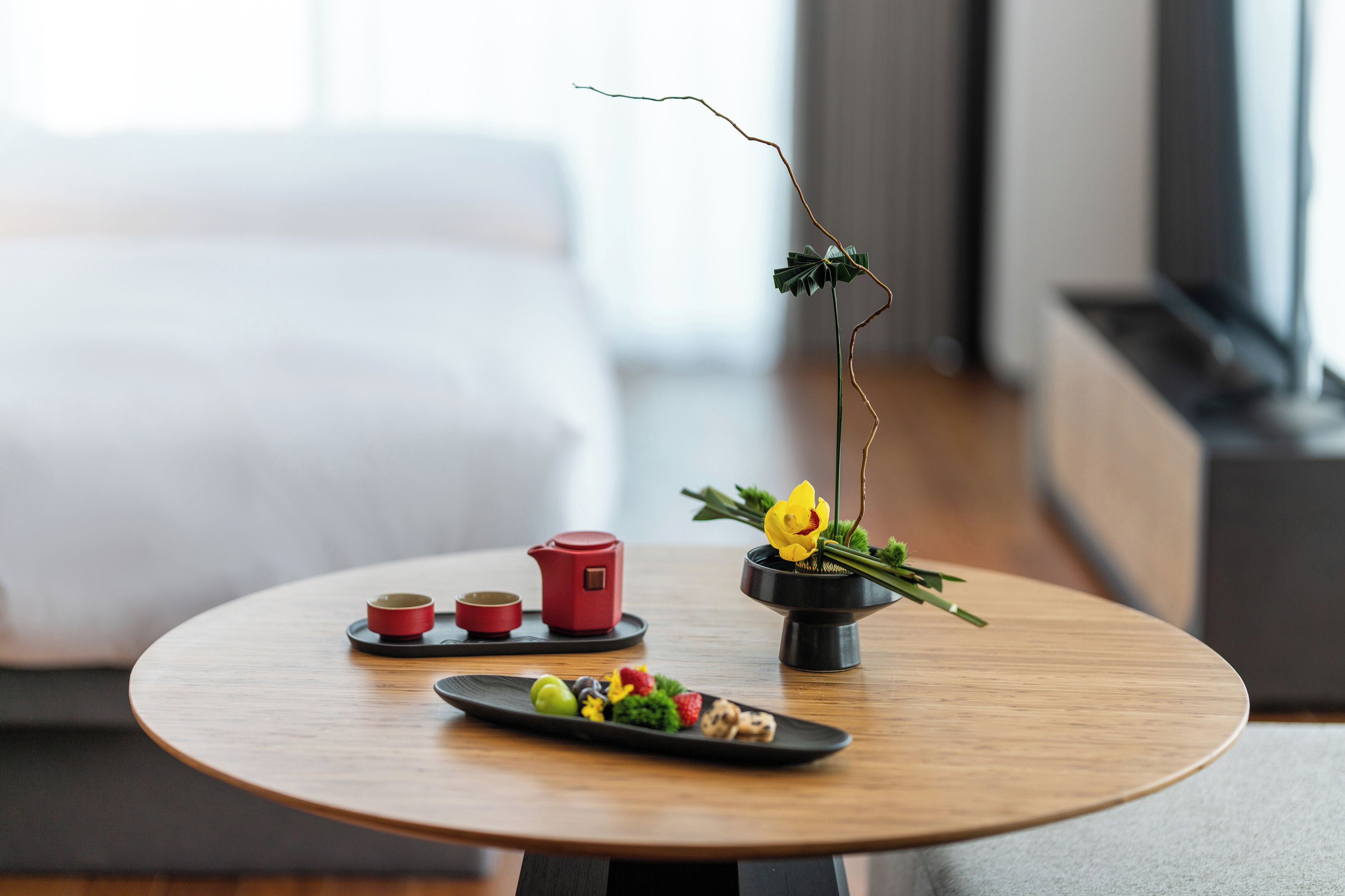 A round wooden table with a tea set, a small vase with a yellow flower, and a plate of food.