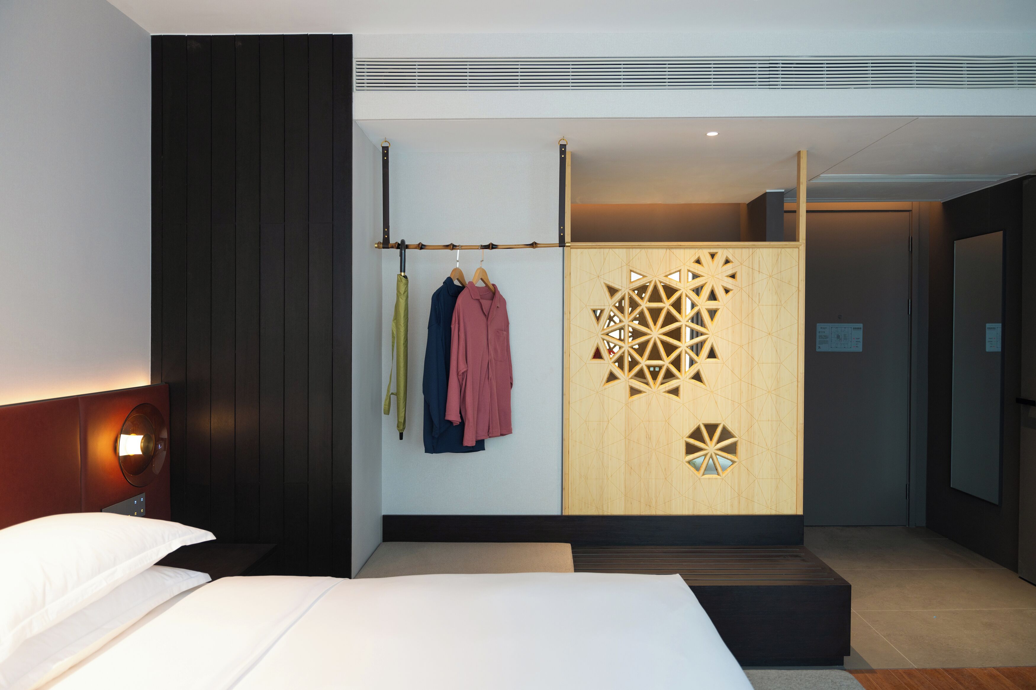 A hotel room with a bed, a wardrobe, a coat rack with clothes, and a decorative wall panel.