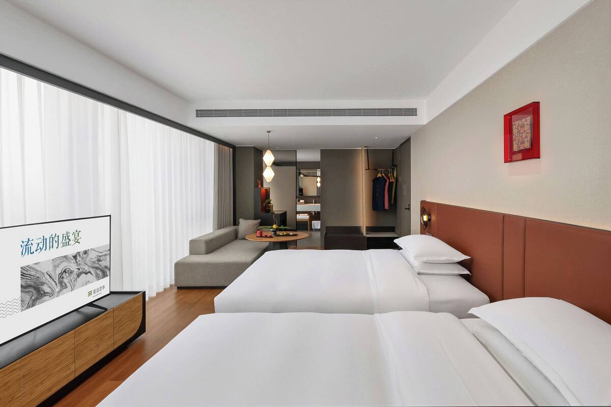 Ein Hotelzimmer mit zwei Betten, einem Fernseher, einem Sofa, einem Esstisch und Blick auf die Stadt.