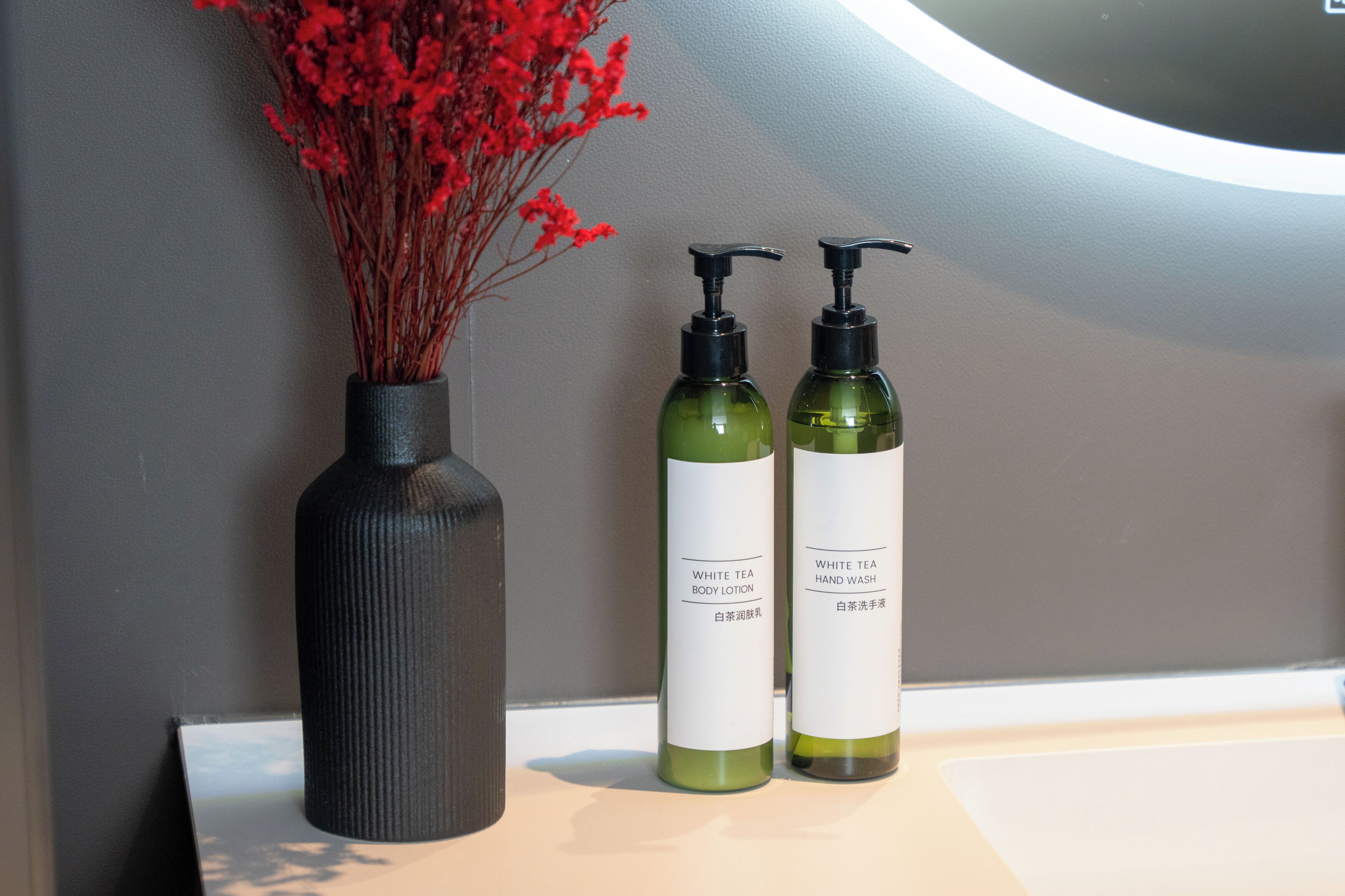 A bathroom vanity with two bottles of white tea body lotion and hand wash, and a vase with red flowers.