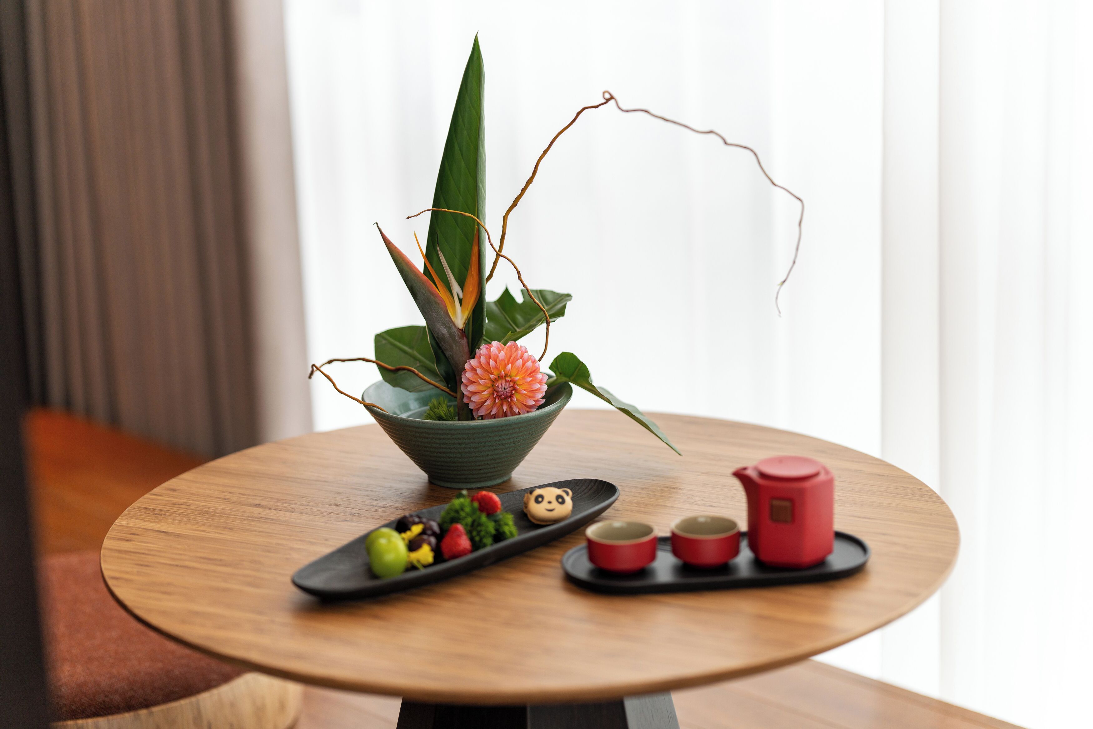 A round wooden table with a green bowl containing a flower and leaves, a black tray with food and a teapot, and two red cups.