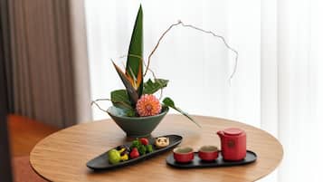 A round wooden table with a green bowl containing a flower and leaves, a black tray with food and a teapot, and two red cups.