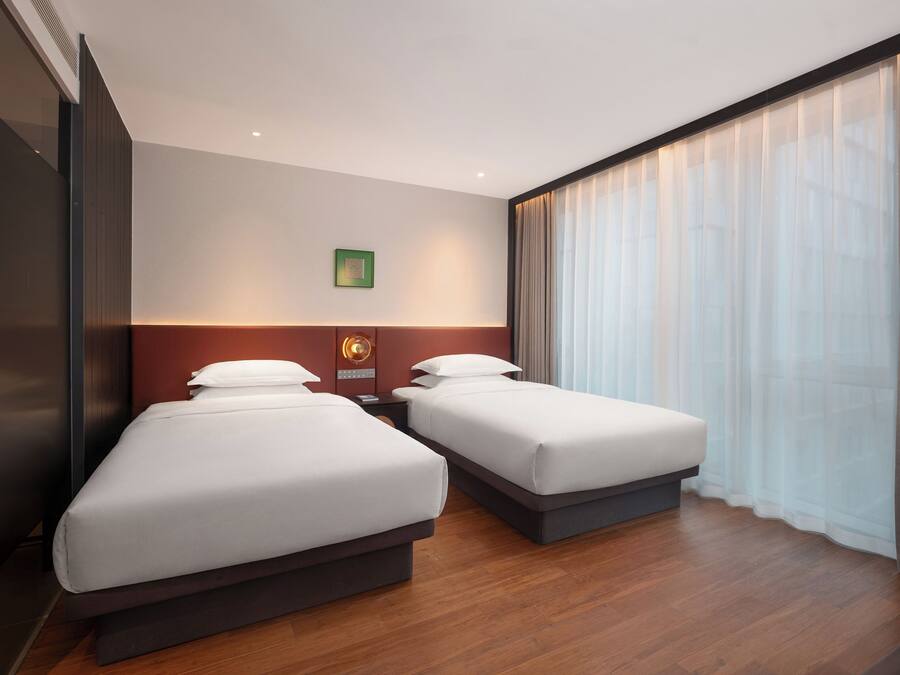 Una habitación de hotel con dos camas, una mesita de noche, un reloj en la pared y un ventanal con cortinas translúcidas.