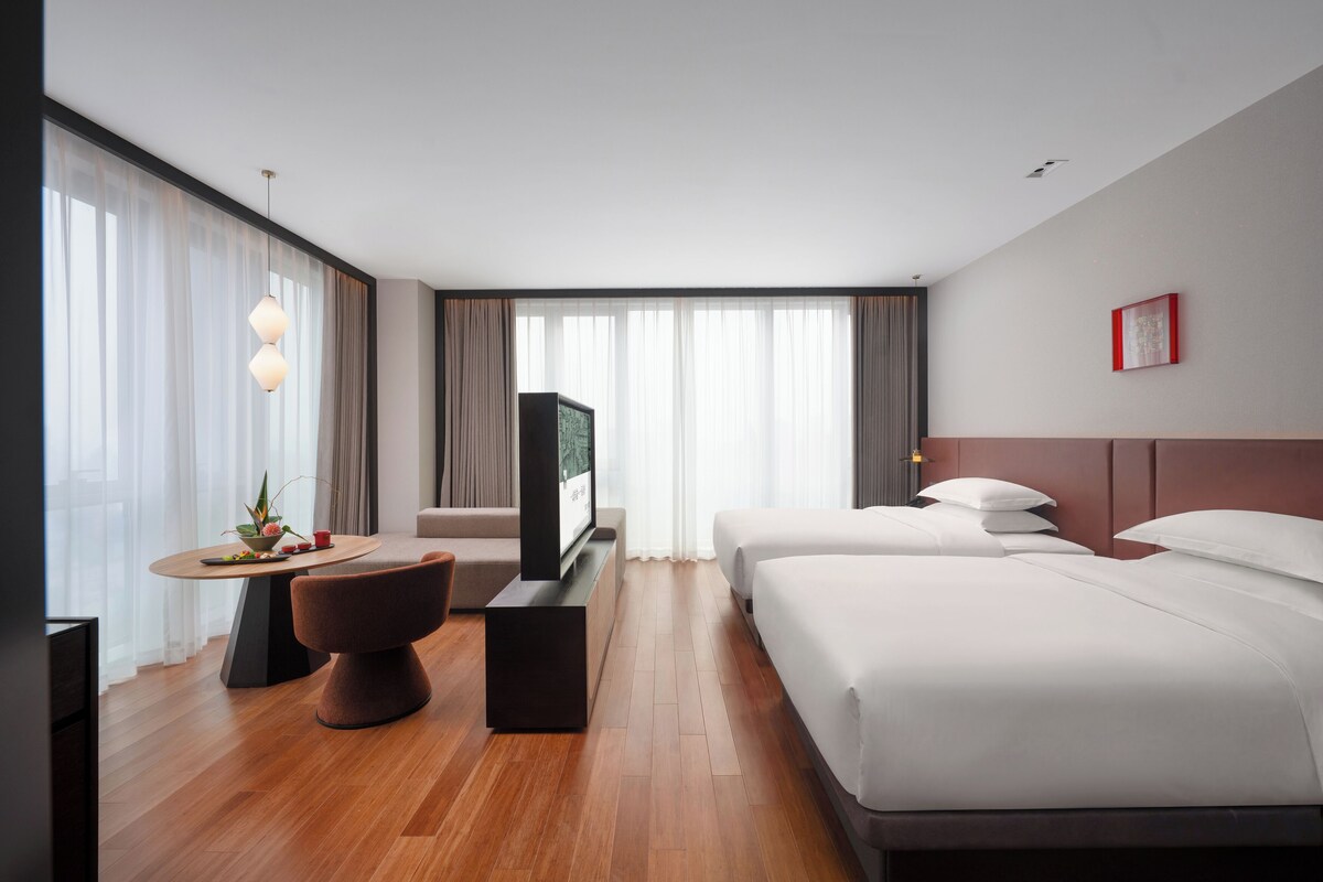 Une chambre d’hôtel avec un plancher en bois, un bureau avec une chaise, deux lits, une télévision et une grande fenêtre avec des rideaux.