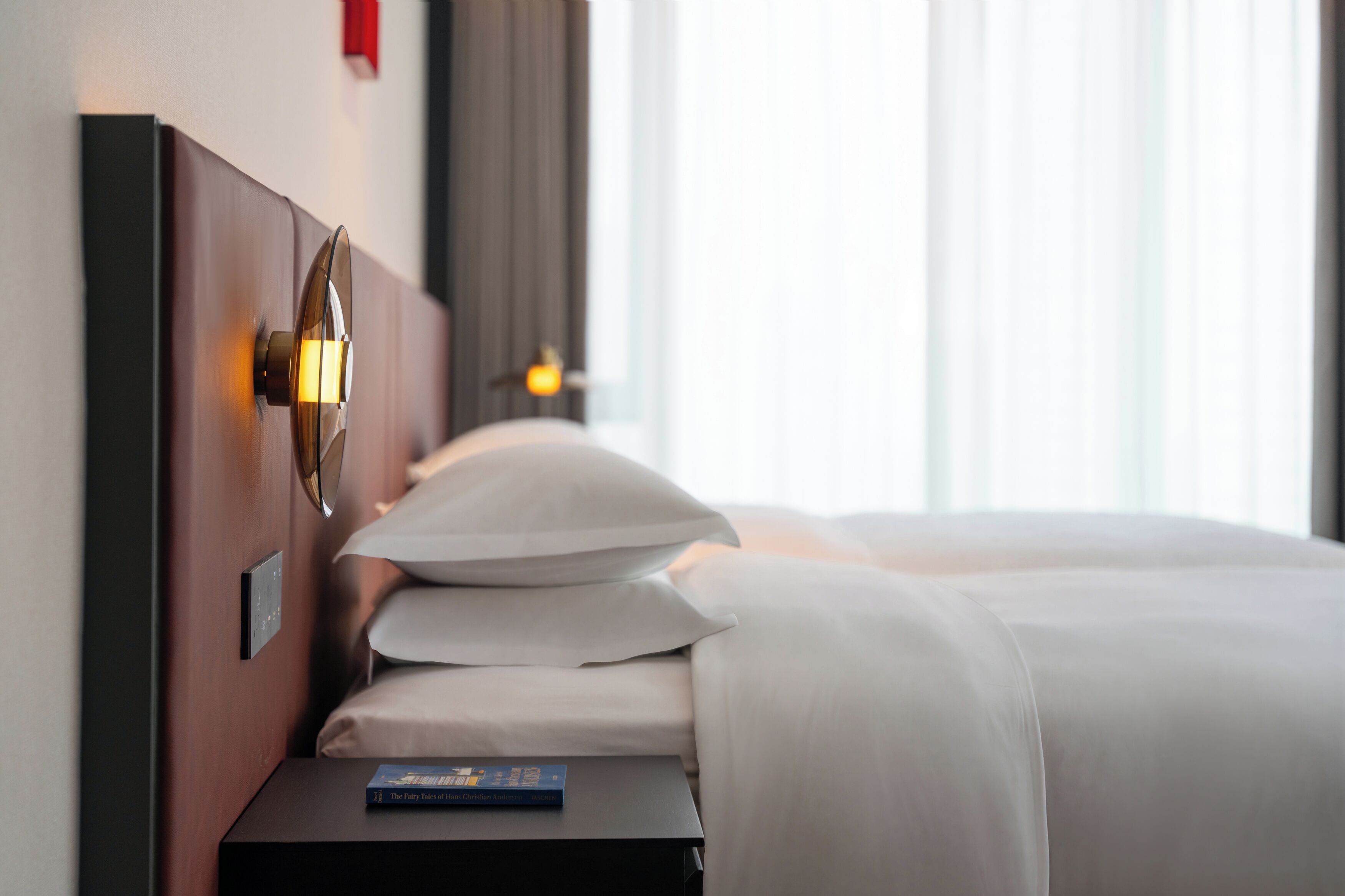Une chambre d’hôtel avec un lit soigneusement fait, une table de chevet sur laquelle repose un livre et un luminaire fixé au mur.
