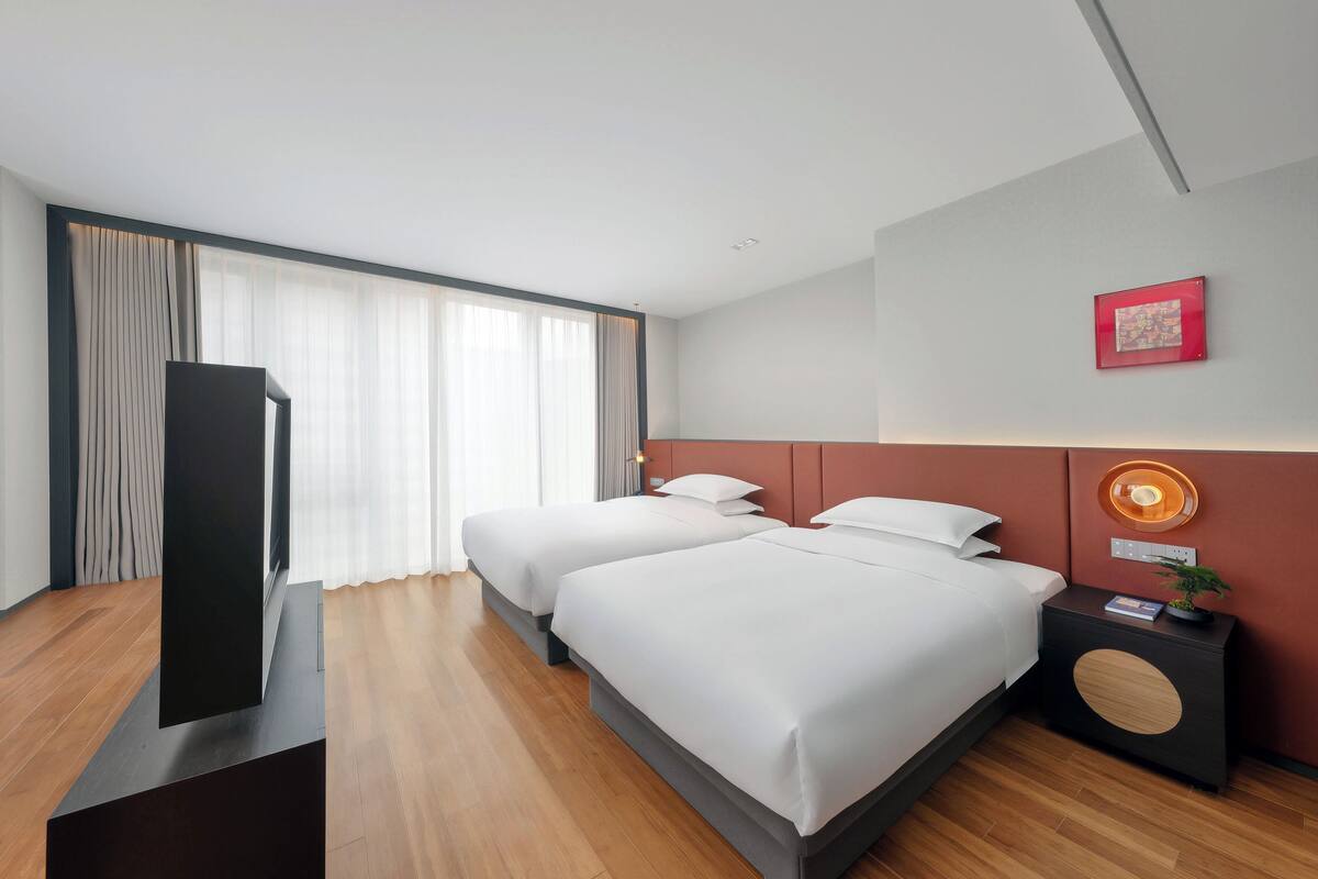 Ein modernes Hotelzimmer mit zwei Betten, einem Flachbildfernseher, einem Nachttisch mit Lampe und einem großen Fenster mit Vorhängen.