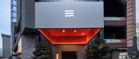 Entrada moderna de un edificio con un logotipo destacado, un toldo de entrada iluminado en rojo y dos árboles en macetas.
