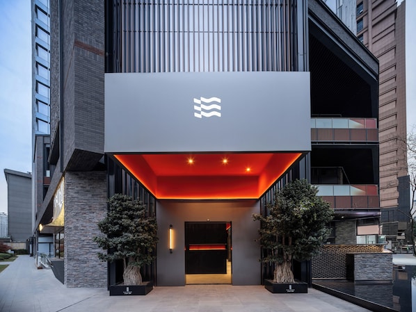 A modern building entrance with a prominent logo, a red-lit entrance canopy, and two potted trees.