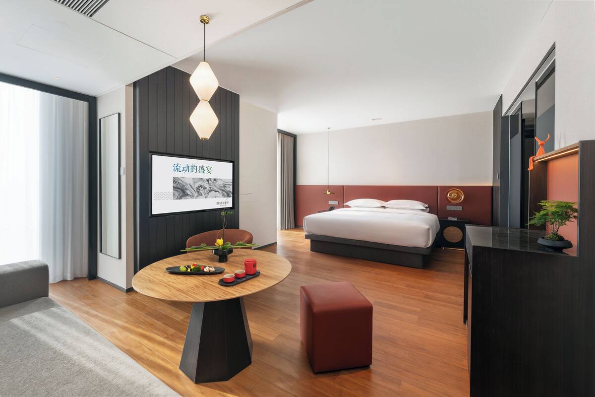 Ein modernes Hotelzimmer mit Holzboden, einem runden Esstisch, einem Bett mit rotem Kopfteil und einem an der Wand montierten Fernseher.