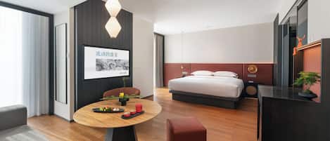 A modern hotel room with a wooden floor, a round dining table, a bed with a red headboard, and a wall-mounted TV.