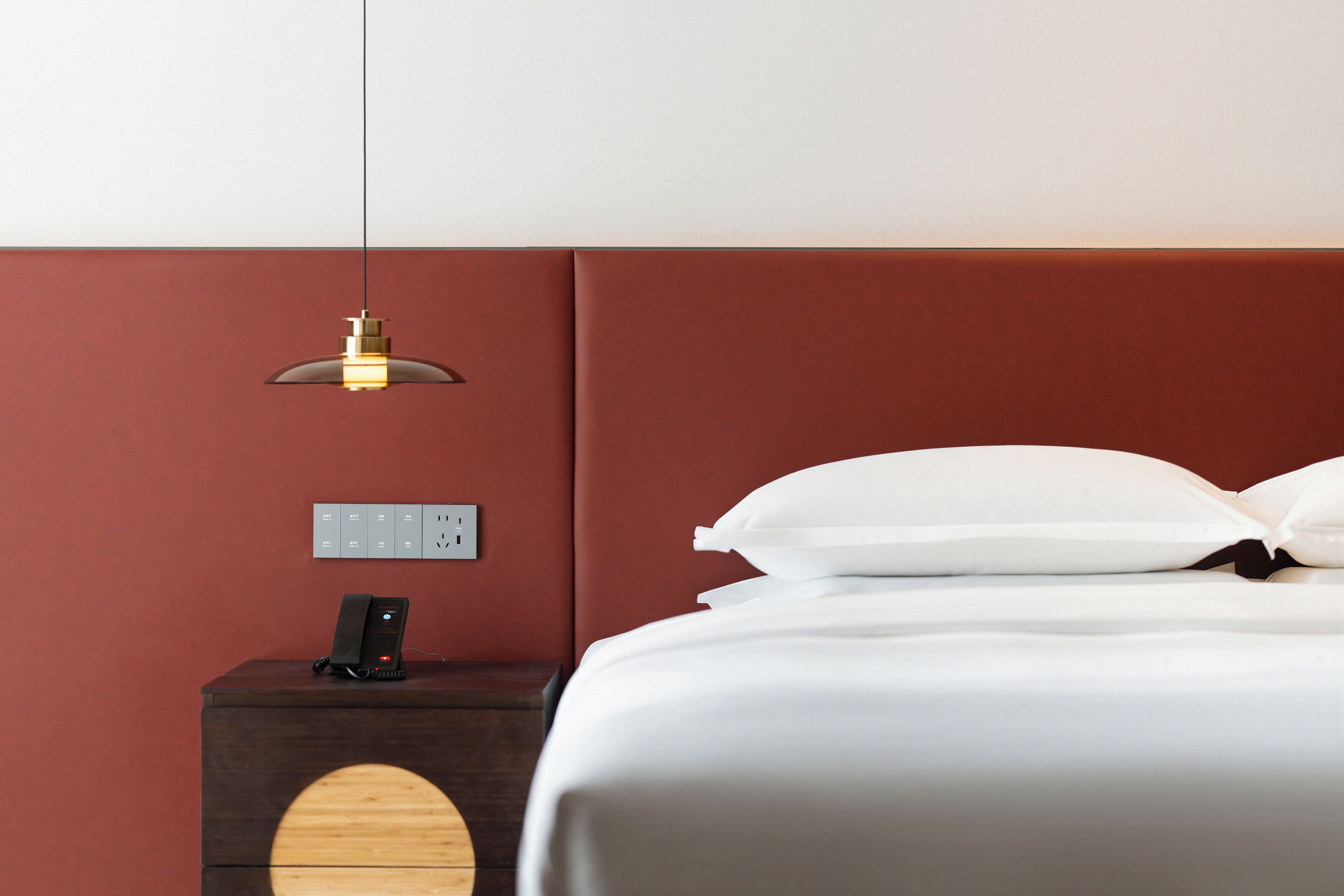 A neatly made bed with white linens, a wooden nightstand with a phone and a lamp, and a wall socket.