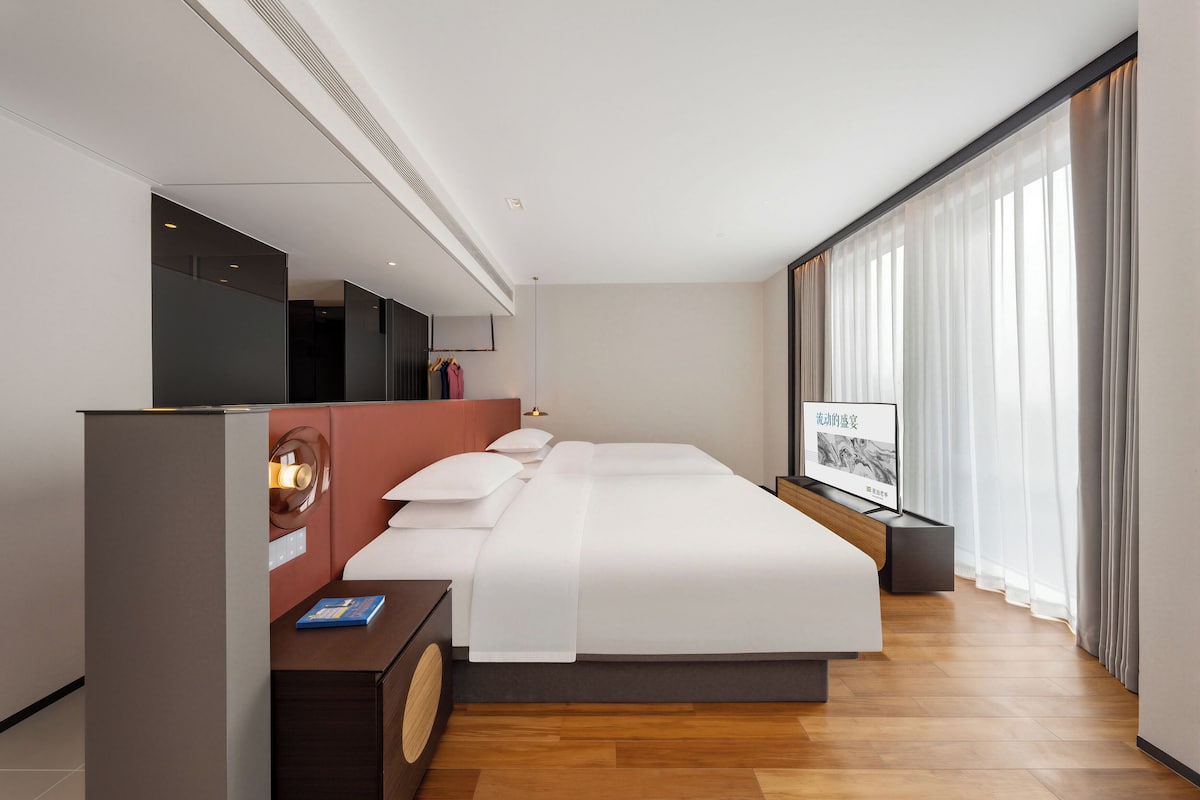 Ein modernes Hotelzimmer mit einem großen Bett, einem Schreibtisch mit Computer, einem Spiegel und einem großen Fenster mit Vorhängen.