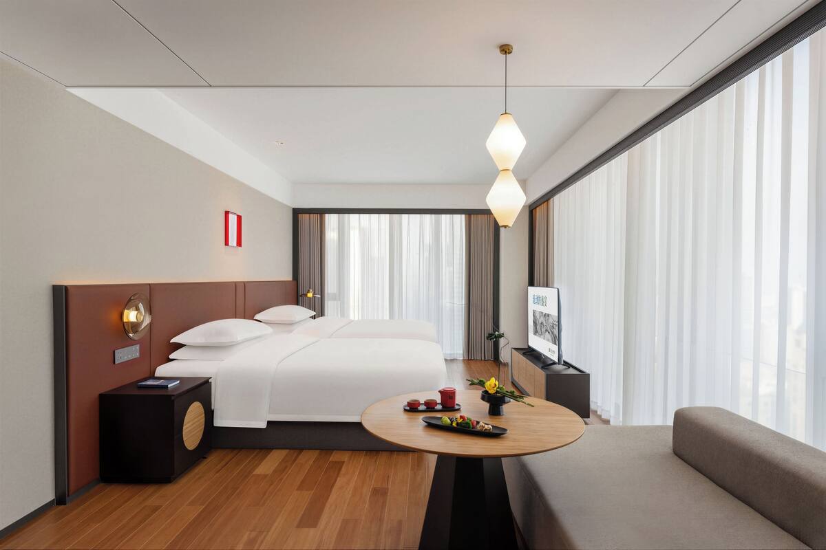 Ein modernes Hotelzimmer mit einem ordentlich bezogenen Bett, einem runden Holztisch mit Blumenvase und einem großen Fenster mit Vorhängen.