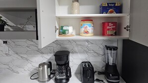 Keittiössä on kahvinkeitin, leivänpaahdin, tehosekoitin sekä hylly, jossa on sokeripurkki, paketti kahvikapseleita ja teepaketti.