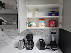 Eine Küche mit Kaffeemaschine, Toaster, Mixer und einem Regal mit Zuckerdose, Kaffeekapseln und einer Teedose.