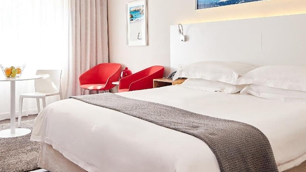 A hotel room with a bed, a desk, a chair, a red chair, a small table with fruit, and a painting on the wall. - The Riviera (Cape Town)