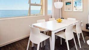 A dining area with a white table and chairs, a large window with a view of the sea, and two framed pictures on the wall. - The Riviera (Cape Town)