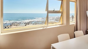 A room with a large window overlooking a rocky beach and ocean. There is a table and two chairs in the room. - The Riviera (Cape Town)