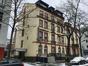 Un hotel a più piani con uno stile architettonico classico, con un cartello 'Hotel West' ben visibile, una recinzione in metallo nero e il terreno coperto di neve.