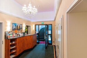 A hotel reception area with a wooden desk, a chandelier, a staircase, and a painting on the wall.